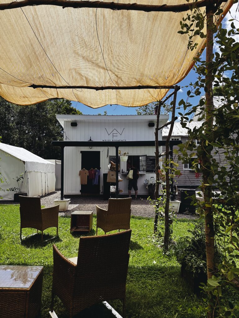 Seated under a cloth canopy with yard chairs nearby and vines growing up the supports. The camera looks on to the small brewhouse and a line of people ordering their next round.