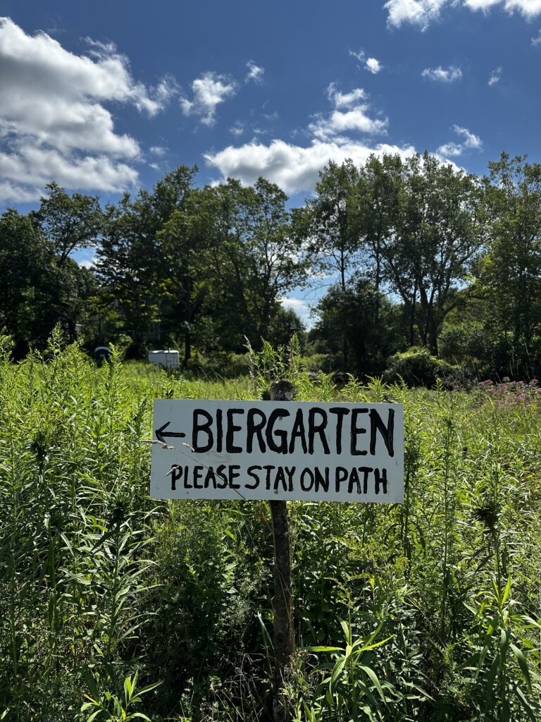 Portrait oriented shot ofa handwritten sign that reads Biergarten. Please Stay on Path with an arrow pointing to the left. It's set in tall grasses and flours with a line of trees behind it.